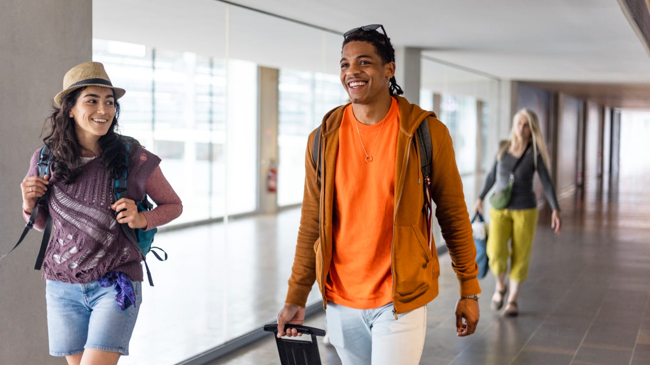 Friends walking through an airport together in Toulouse, France. They are heading to their departure area while carrying luggage and smiling.