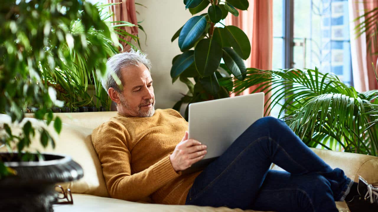 A middle-aged man sits on a couch surrounded by plants, looking at a laptop.
