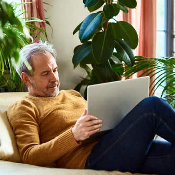 A middle-aged man sits on a couch surrounded by plants, looking at a laptop.