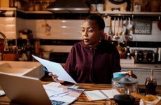 A Black woman concentrates while doing finances; she's in front of a countertop with paperwork and a laptop sitting on it.