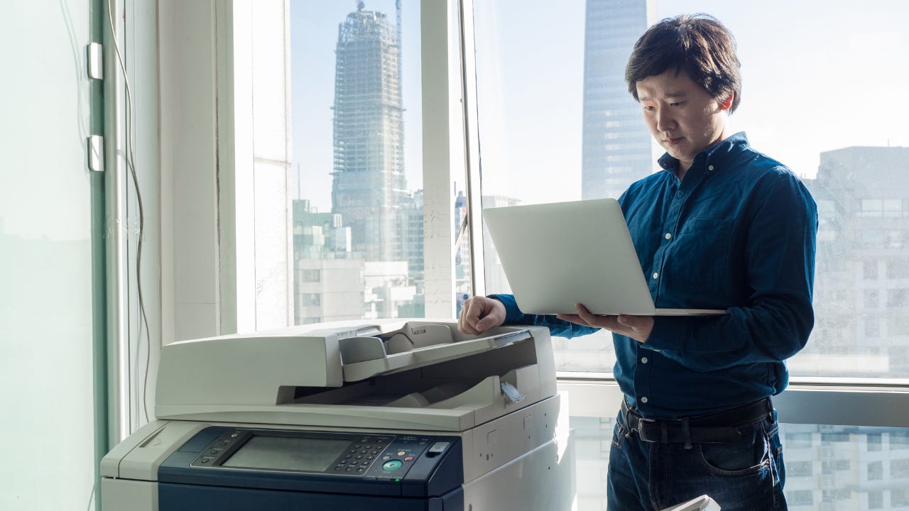 An entrepreneur stands by the printer in his office, holding his laptop.