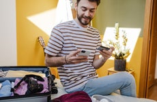 Young smiling man in a striped t-shirt using mobile phone and holding credit card at bedroom. Getting ready for a vacation.