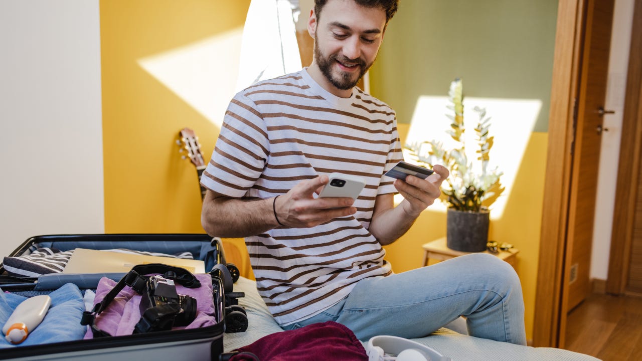 Young smiling man in a striped t-shirt using mobile phone and holding credit card at bedroom. Getting ready for a vacation.