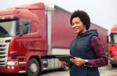 A business owner stands in front of her semi-trucks while holding a tablet.