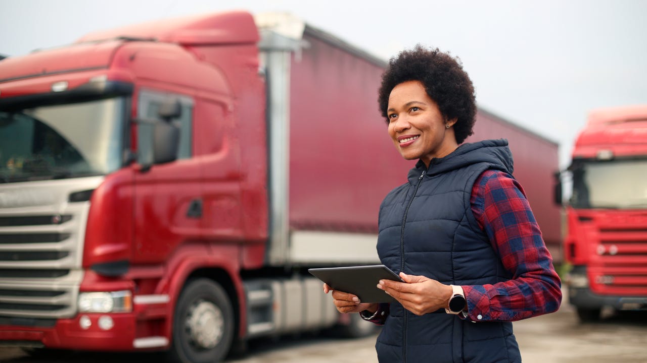 A business owner stands in front of her semi-trucks while holding a tablet.