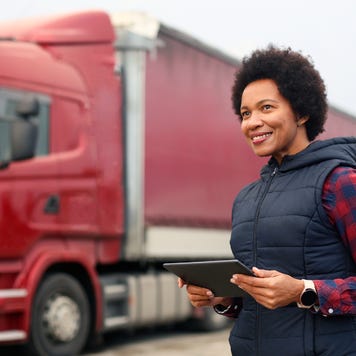 A business owner stands in front of her semi-trucks while holding a tablet.