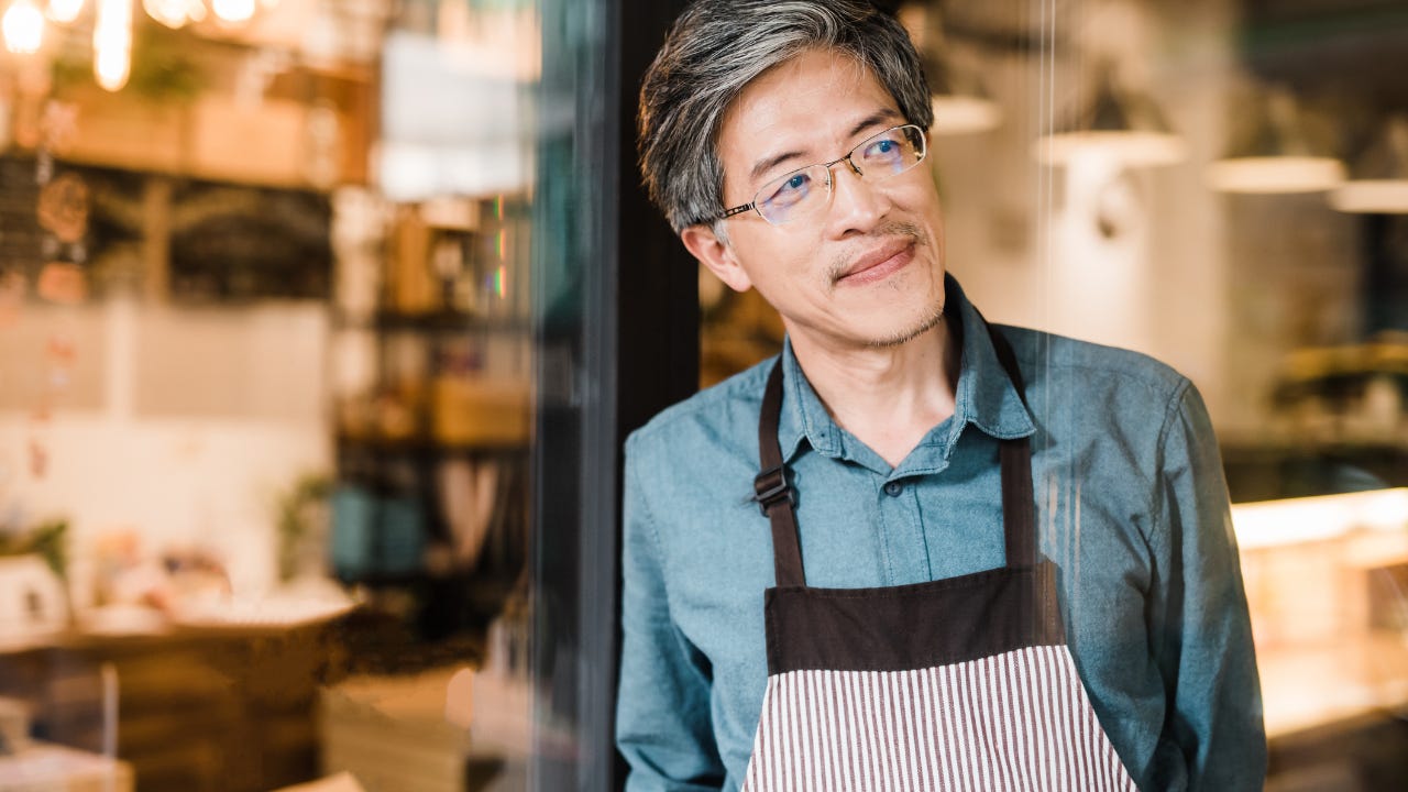 A small business owner smiles as he leans against the doorway of his business, looking down the street.