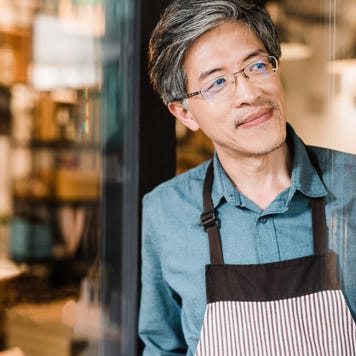 A small business owner smiles as he leans against the doorway of his business, looking down the street.