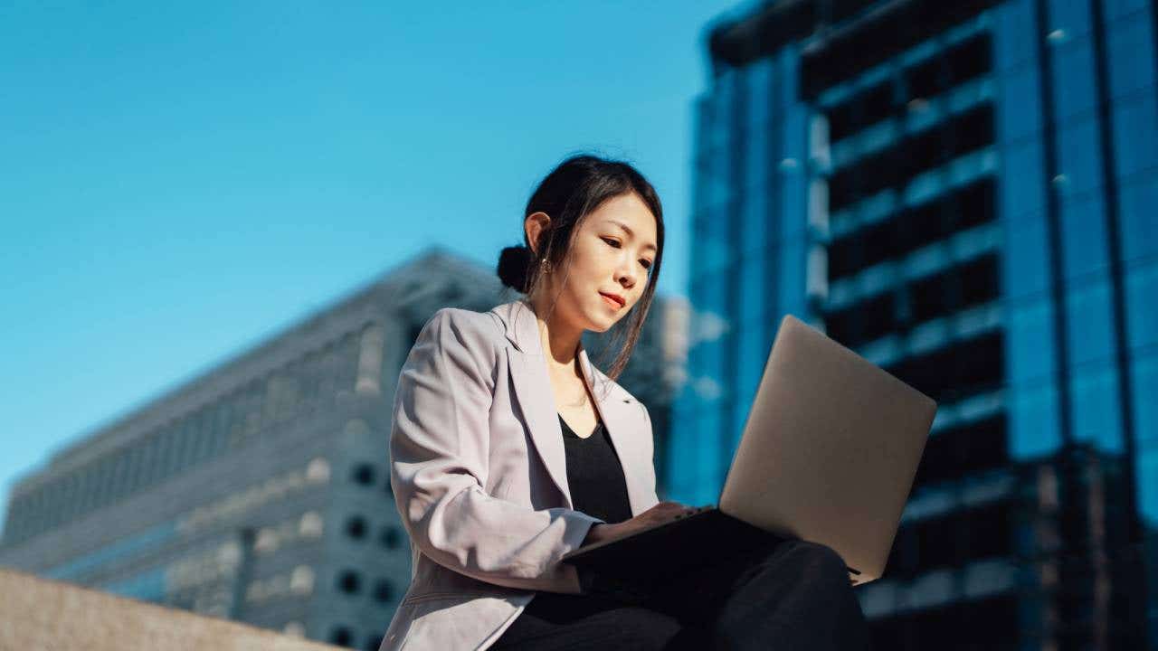 Young businesswoman working on laptop, sitting against financial buildings in the city
