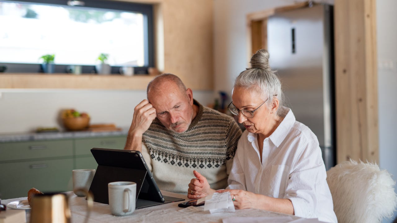 Senior couple sitting at the kitchen table looking at digital tablet and recalculating their expenses.