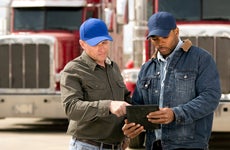 Two semi-truck drivers stand in front of their rigs, looking at a tablet.