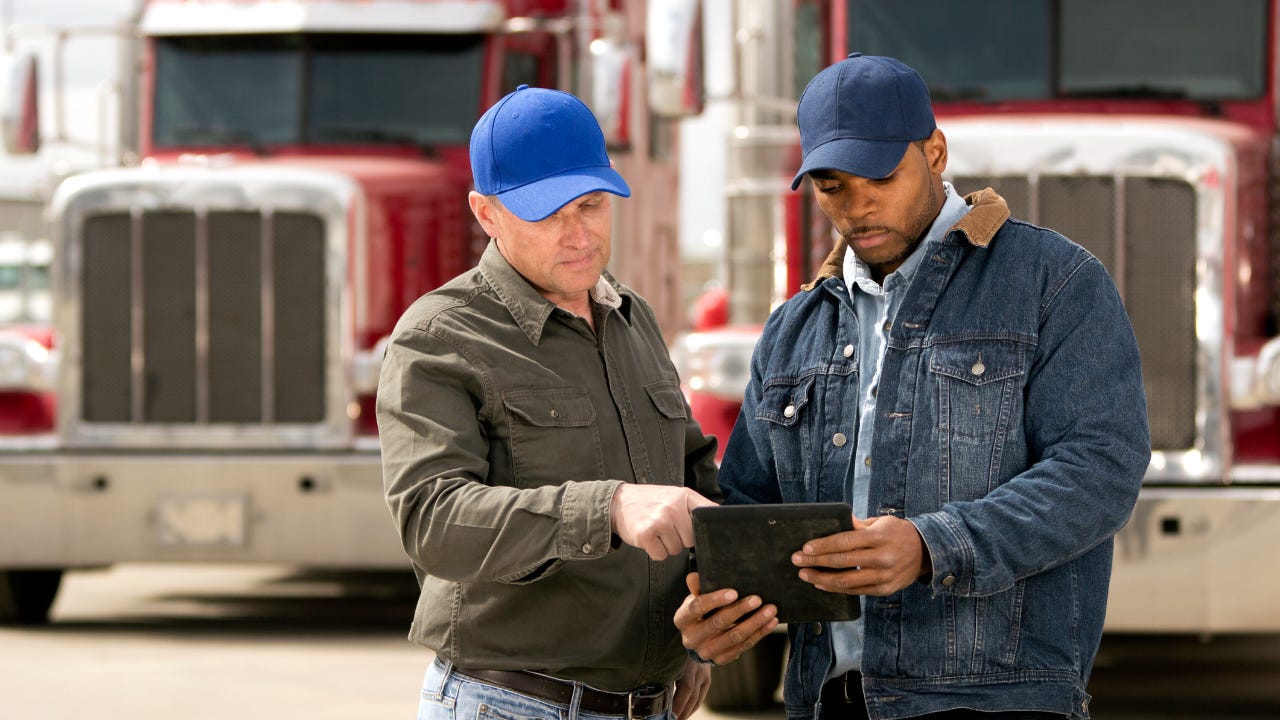 Two semi-truck drivers stand in front of their rigs, looking at a tablet.