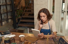 A young woman works at her table, talking on the phone while looking at her laptop.
