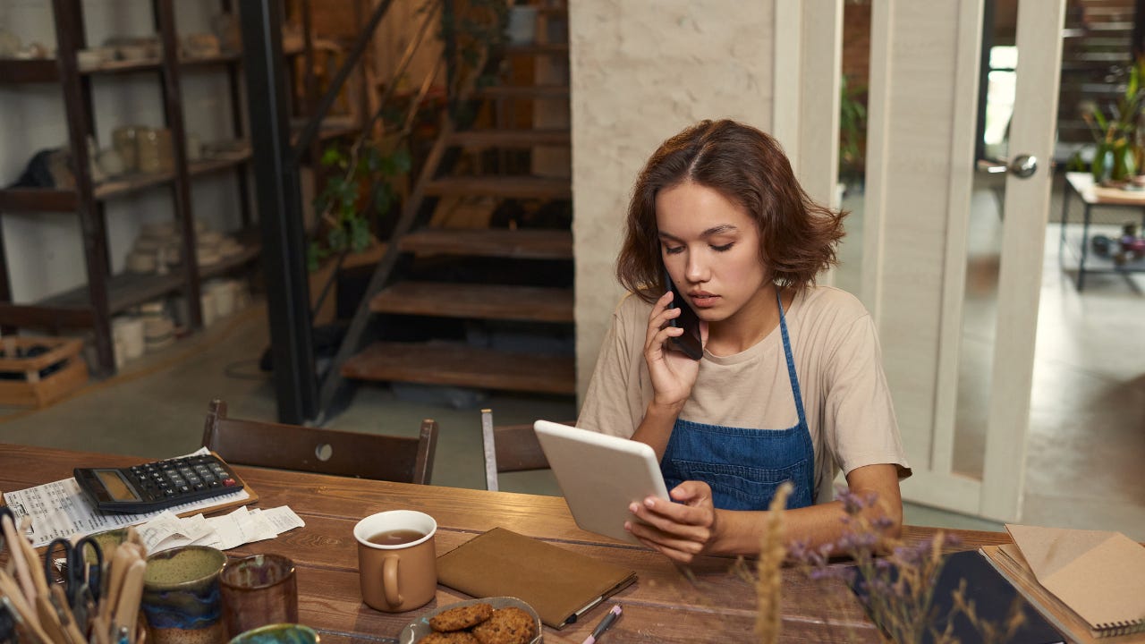 A young woman works at her table, talking on the phone while looking at her laptop.