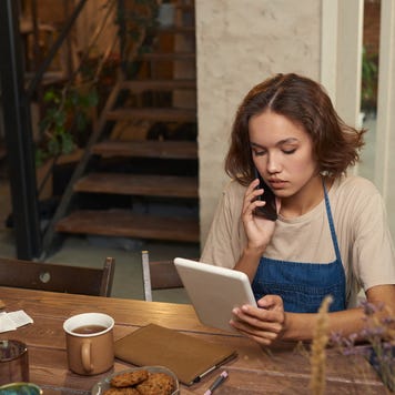 A young woman works at her table, talking on the phone while looking at her laptop.