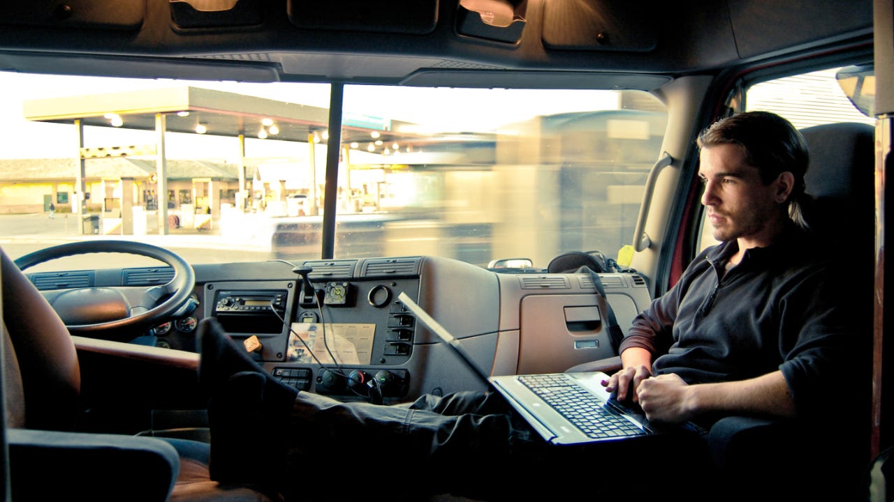 A truck driver relaxes in his cab, looking at his laptop.