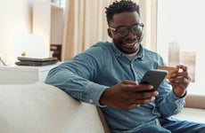 Shot of a young man holding his credit card while using a smartphone at home. Cheerful young man doing online shopping while using his cellphone and credit card inside of an apartment during the day.