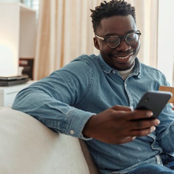 Shot of a young man holding his credit card while using a smartphone at home. Cheerful young man doing online shopping while using his cellphone and credit card inside of an apartment during the day.
