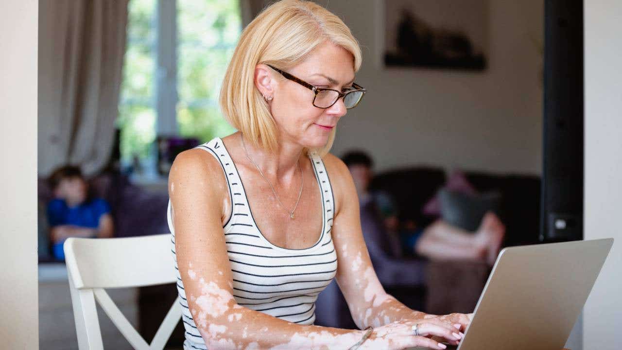 Blonde woman sitting her dining table at home with a laptop open in front of her as she works