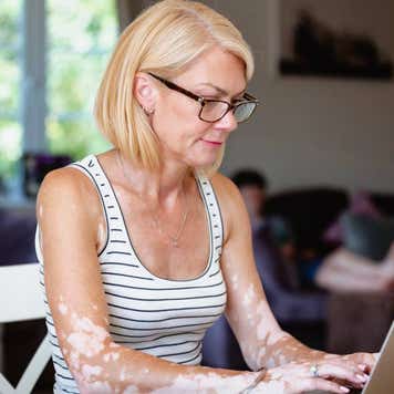 Blonde woman sitting her dining table at home with a laptop open in front of her as she works