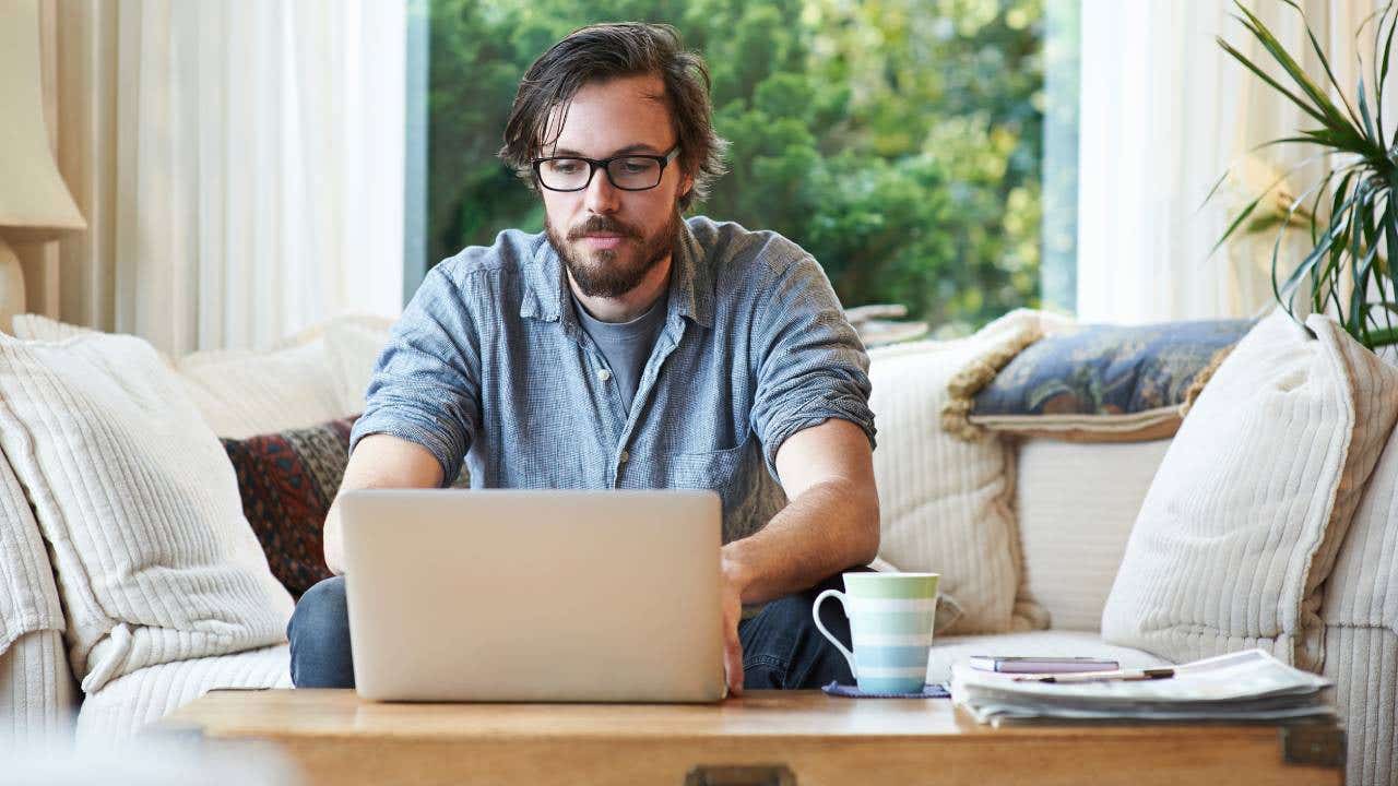A man sits on the sofa in the living room of his home and uses a laptop.