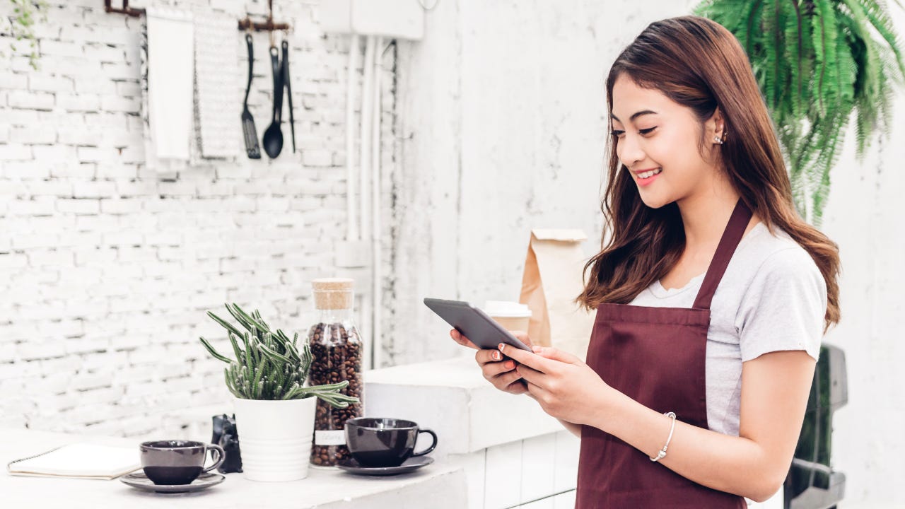 A young business owner, wearing an apron, smiles as she looks at her tablet.