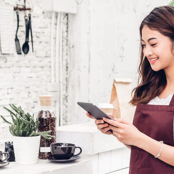 A young business owner, wearing an apron, smiles as she looks at her tablet.