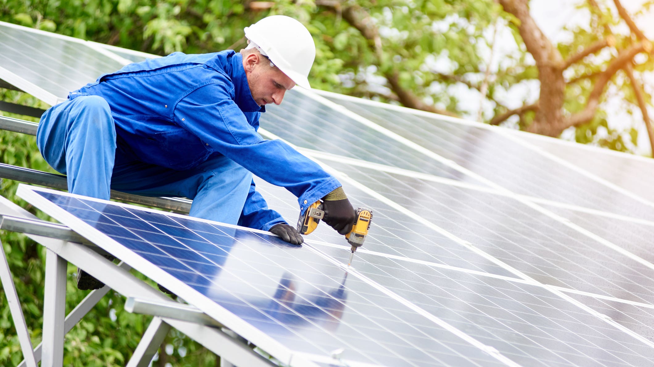 Young construction worker connects photo voltaic panel to solar system using screwdriver on bright sunny day.