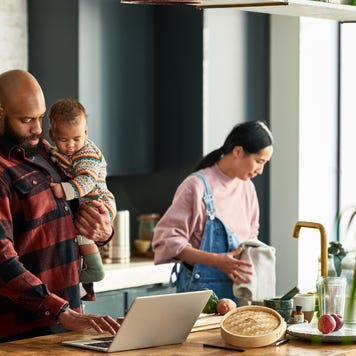 A mother prepares a meal in the kitchen as father holds baby boy and uses a laptop.