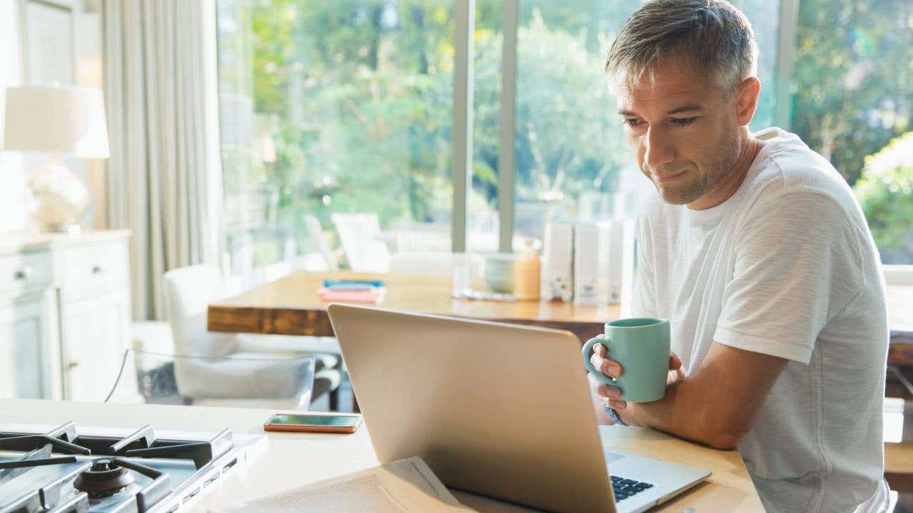 Man drinking coffee and working at laptop in kitchen