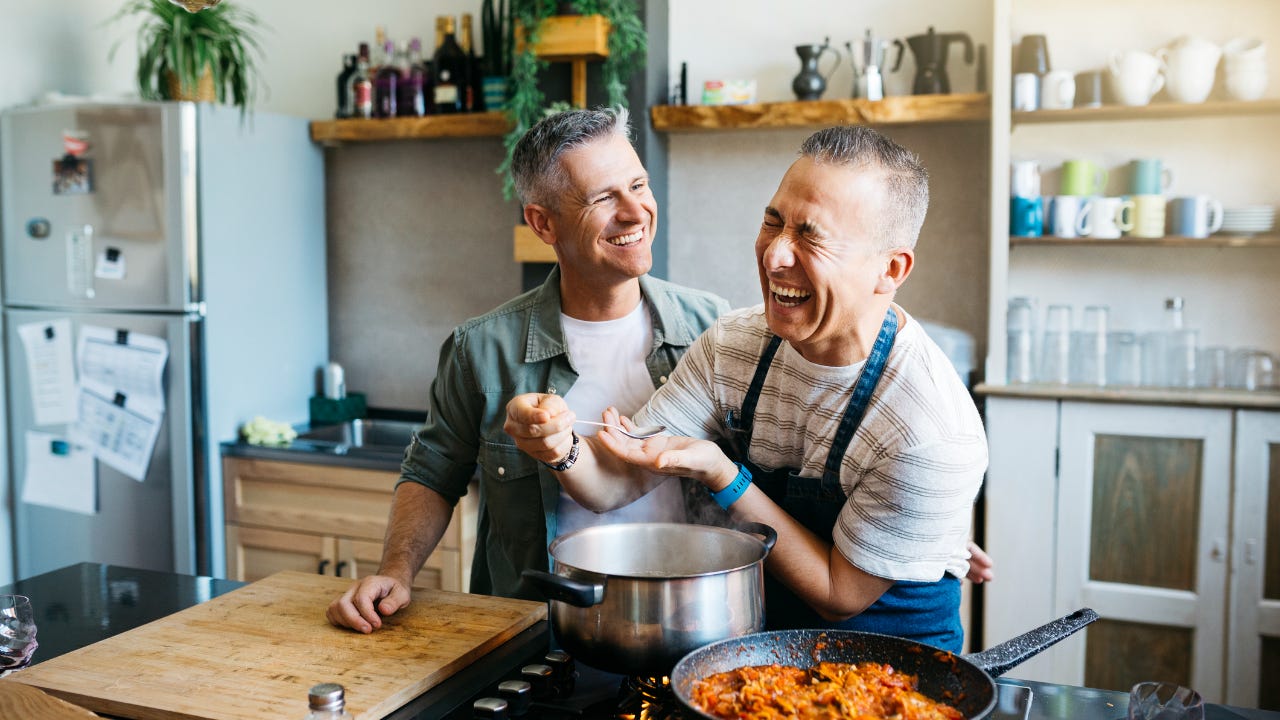 A chef prepares a dish in his kitchen while laughing with his partner.