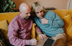 A gay couple sit on a yellow sofa and use a laptop together. One man holds a credit card. They are relaxed and content as they have a lighthearted discussion. Wall provides a space for copy.