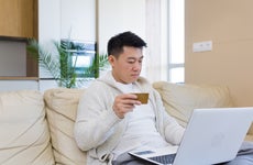 young asian man shopping online in internet store using laptop computer and credit card. male enters data of paying for services or tickets in application sitting on couch at home living room