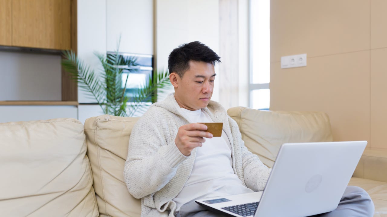 young asian man shopping online in internet store using laptop computer and credit card. male enters data of paying for services or tickets in application sitting on couch at home living room