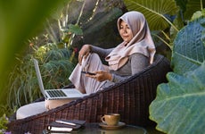 Asian woman wearing Hijab working from home using laptop and mobile phone, sitting in garden furniture surrounded by tropical plants.