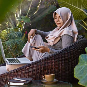 Asian woman wearing Hijab working from home using laptop and mobile phone, sitting in garden furniture surrounded by tropical plants.