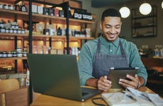 A shop owner wearing an apron smiles as he works on a tablet and laptop in his shop.
