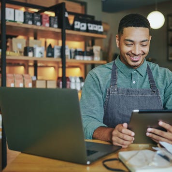 A shop owner wearing an apron smiles as he works on a tablet and laptop in his shop.
