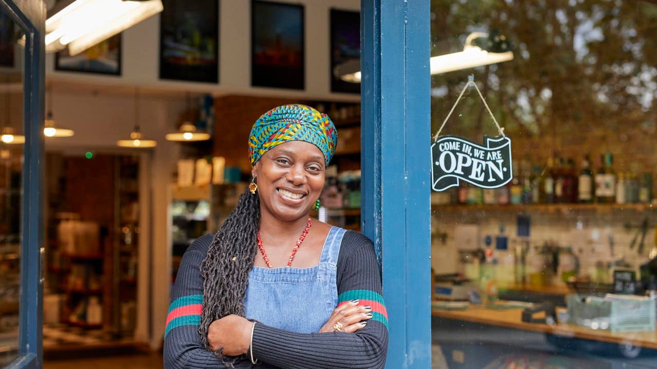 Portrait of black female shop owner in shop door way. She is smiling, looking at the camera. She has braided hair.
