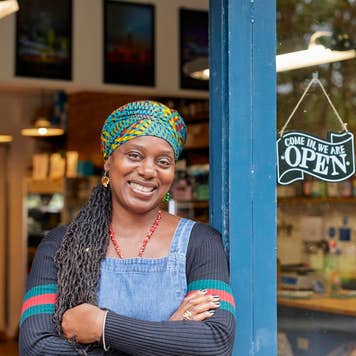 Portrait of black female shop owner in shop door way. She is smiling, looking at the camera. She has braided hair.