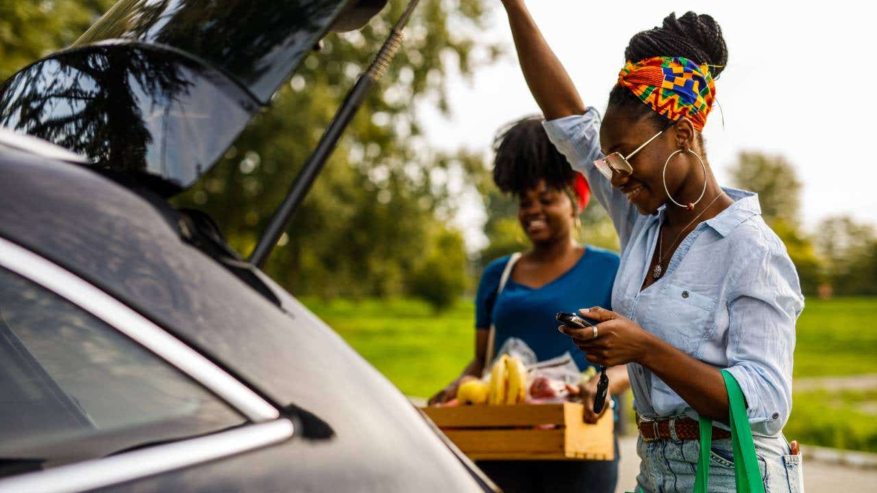 Candid shot of young Black woman opening trunk on her car to load a reusable shopping bag and a crate with groceries with a help of her friend.