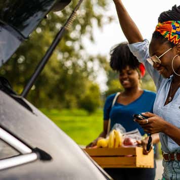 Candid shot of young Black woman opening trunk on her car to load a reusable shopping bag and a crate with groceries with a help of her friend.