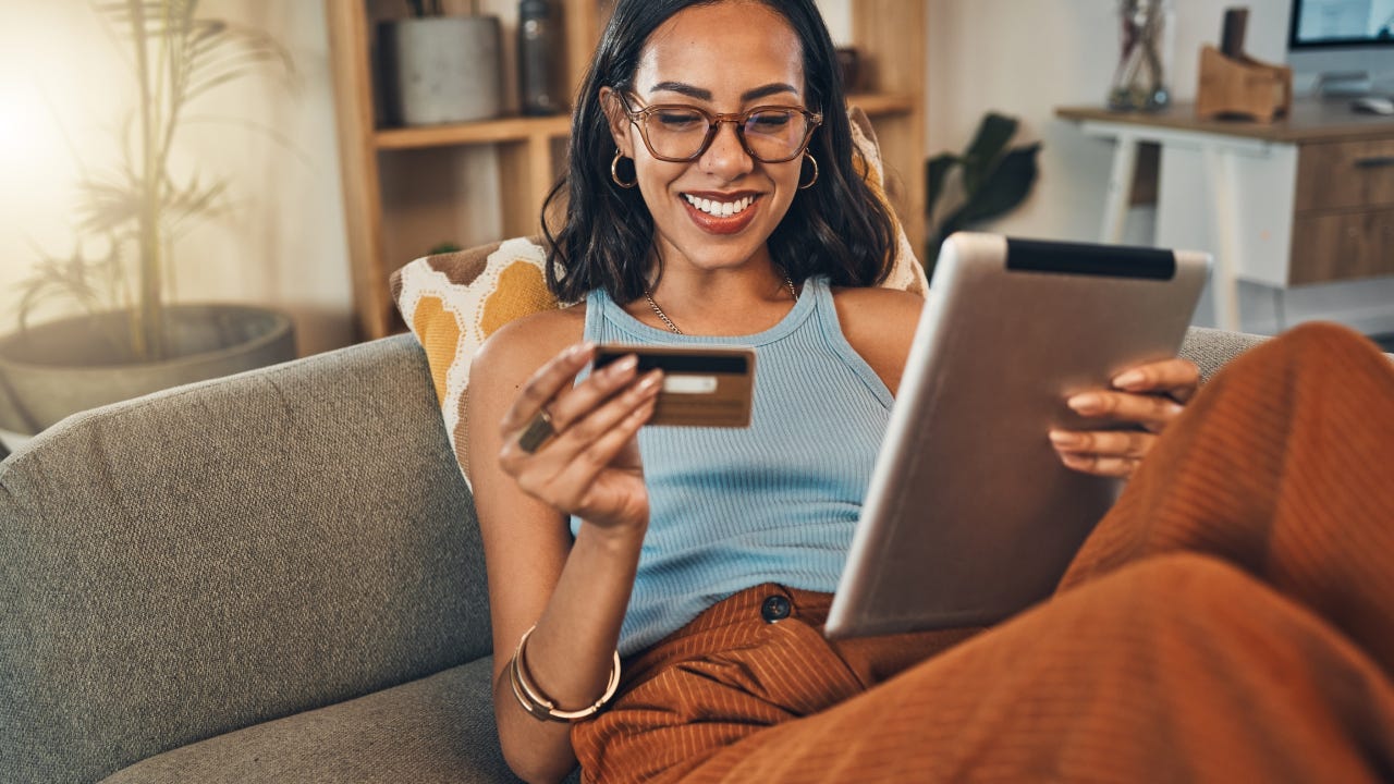 Smiling mixed race woman using credit card for ecommerce on digital tablet at home. Happy hispanic sitting alone on living room sofa, using technology for ebanking. Relaxing, ordering, buying online