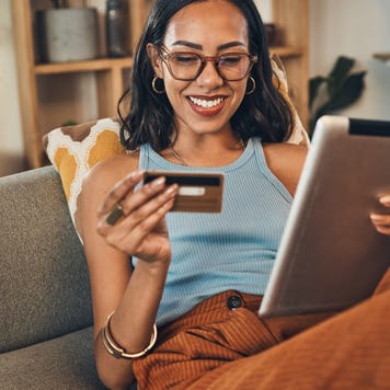 Smiling mixed race woman using credit card for ecommerce on digital tablet at home. Happy hispanic sitting alone on living room sofa, using technology for ebanking. Relaxing, ordering, buying online