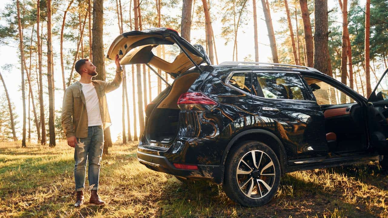 Man in jeans is outdoors in the forest with his black colored automobile