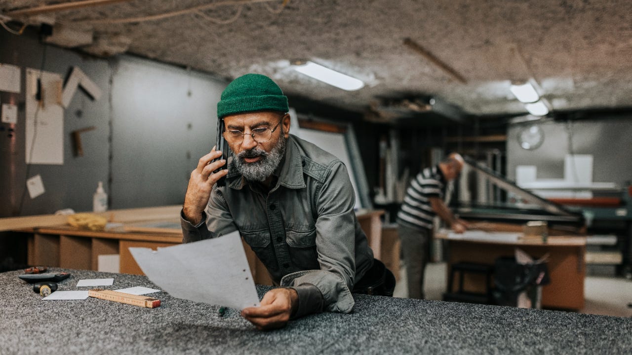 Man with beard looks over paperwork while talking on the phone in a workshop.