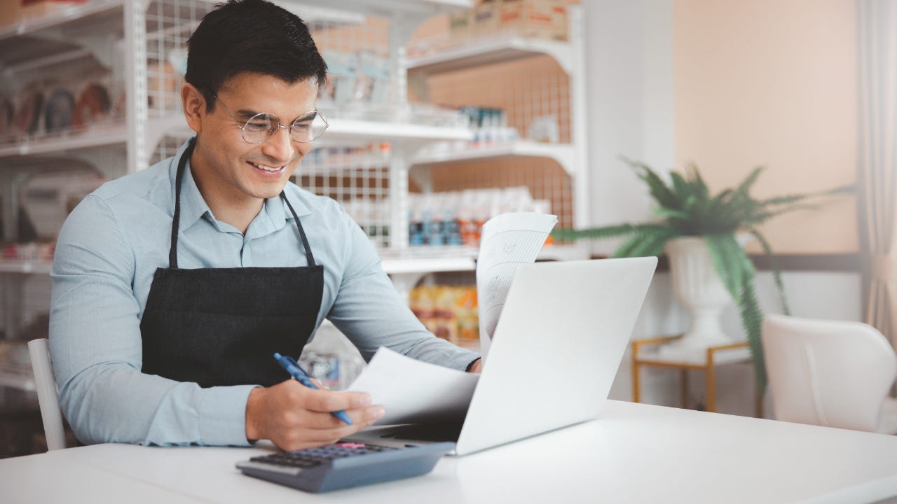 Male business owner in glasses using a laptop while reviewing documents.