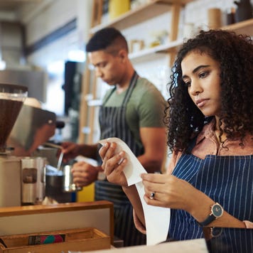 A young business owner stands behind the counter of her coffee shop, looking discouragingly at the day’s receipts.