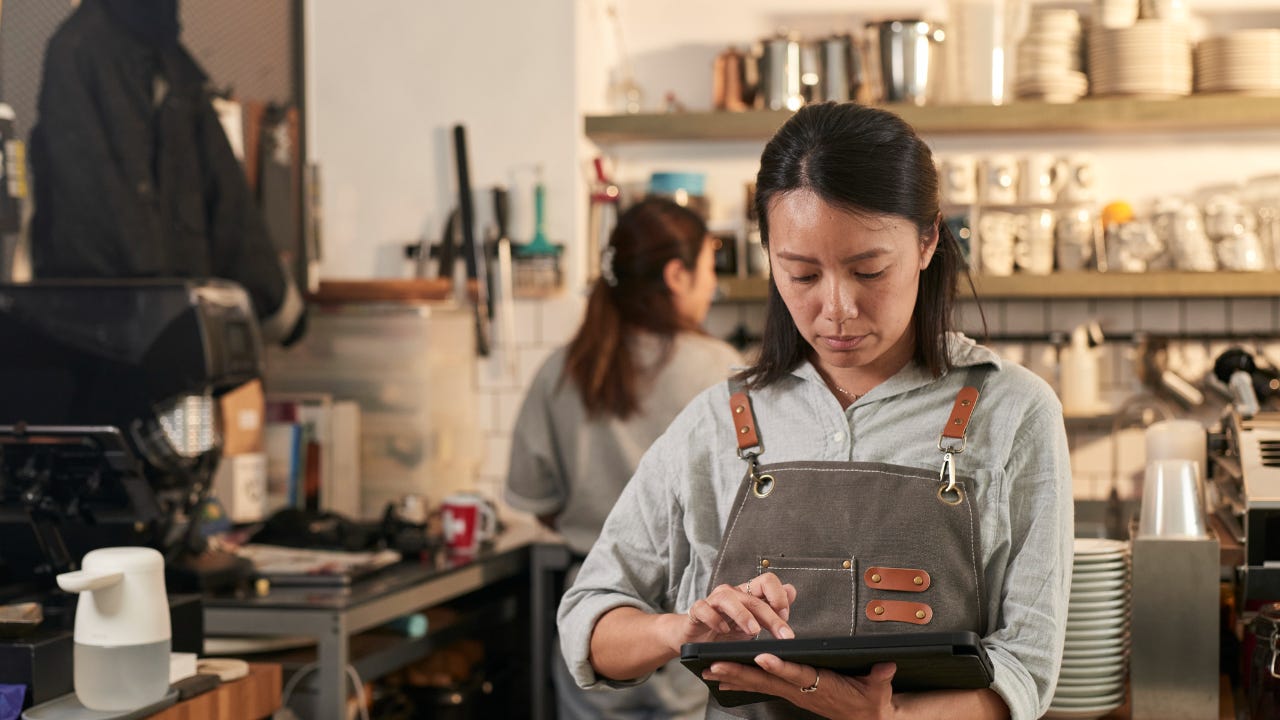 A cafe owner in coffee shop and wearing an apron holds a digital tablet.