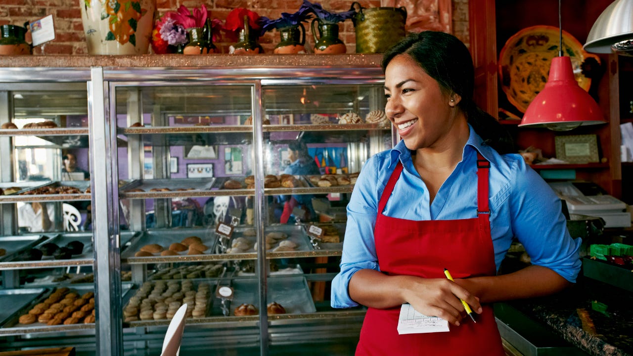 A bakery owner, wearing a red apron, smiles as she stands at the counter.
